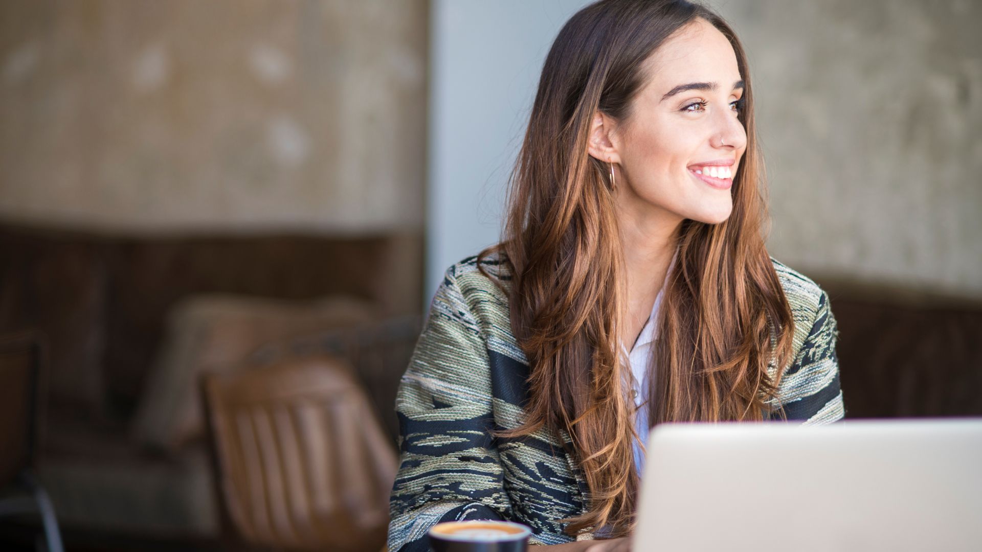 A woman with long brown hair smiles while looking to the side, sitting at a desk with a laptop and a cup of coffee. The screen shows a list of therapy business names.